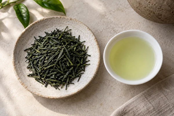 Dunkle Gyokuro-Nadeln neben einer kleinen Tasse helljadefarbenem Tee
