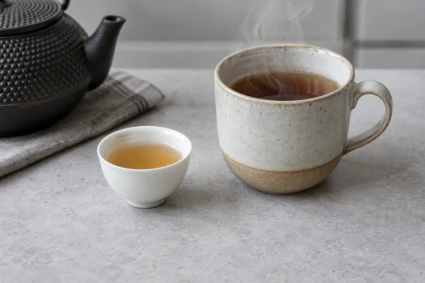 Eine kleine Probiertasse neben einer größeren dampfenden Teetasse