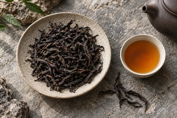 Lange dunkle Wuyi-Oolongblätter neben bernsteinfarbenem Tee auf grauem Stein